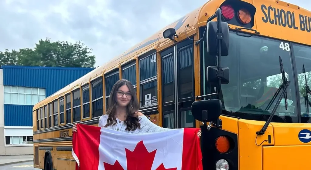 Estudiante sonriente sosteniendo la bandera canadiense frente a un autobús escolar amarillo durante su año escolar en Canadá.